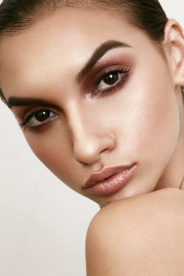 Close-up beauty portrait of a woman with dramatic burgundy eye makeup and glossy nude lips against a plain background.