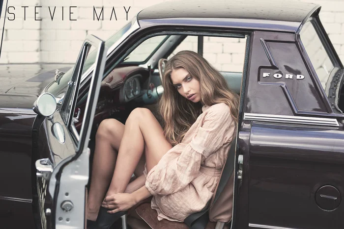 Model wearing a light-coloured dress seated inside a vintage dark-coloured Ford vehicle with the door open.