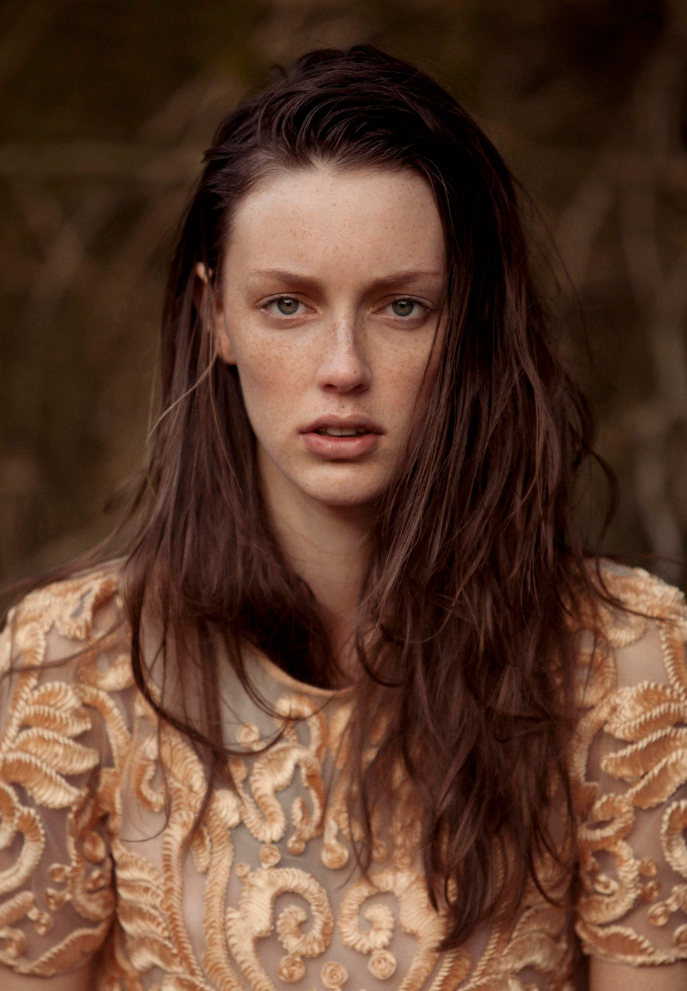 Portrait of a woman with long brown hair and freckles wearing a beige embroidered dress against a dark woodland background