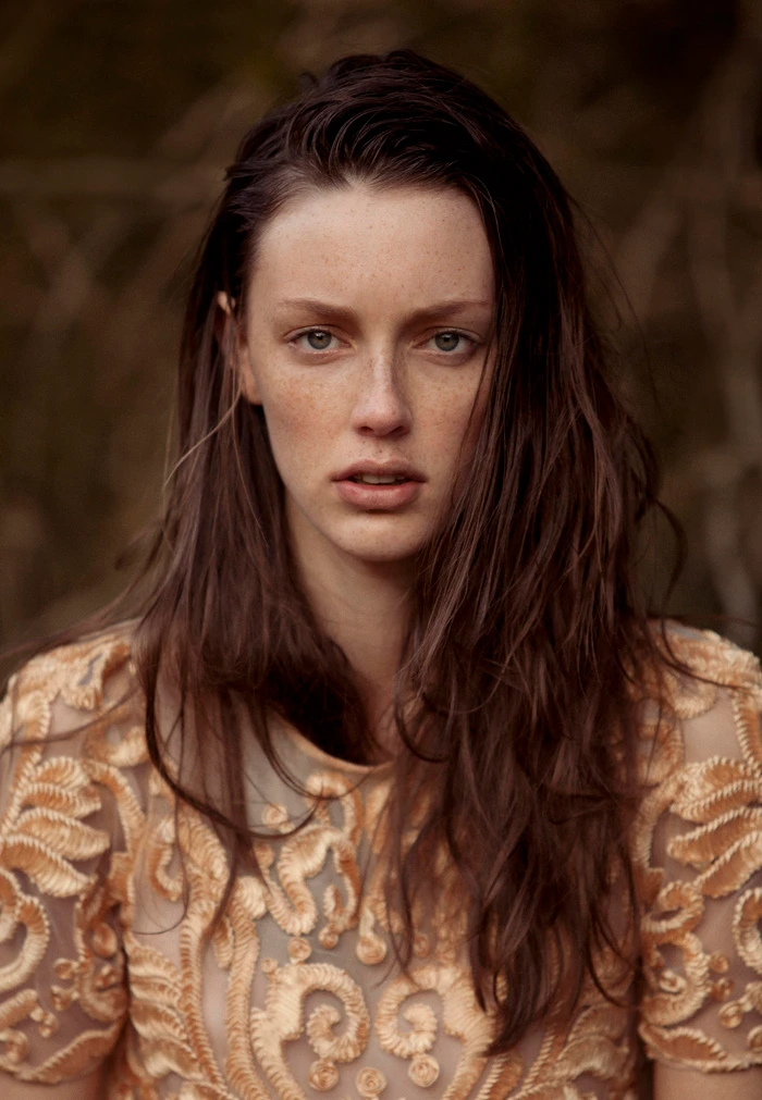 Portrait of a woman with long brown hair and freckles wearing a beige embroidered dress against a dark woodland background