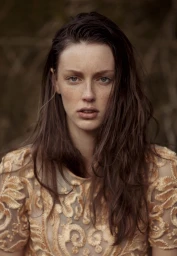 Portrait of a woman with long brown hair and freckles wearing a beige embroidered dress against a dark woodland background