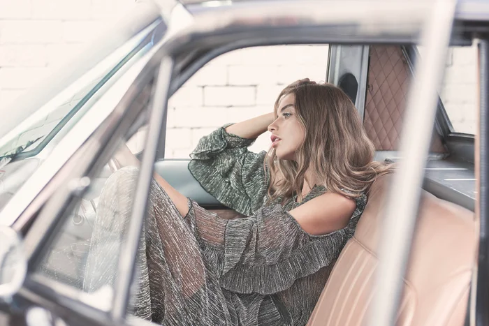 Woman with long wavy hair wearing a metallic green dress seated inside a vintage car with tan leather interior