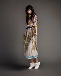 Model with dark tousled hair and blue eye makeup wearing a patterned skirt and white sneakers against a grey backdrop