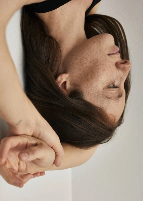 Close-up portrait of a freckled woman lying down with hands behind her head against a light background in a brightly lit, eye-level shot.