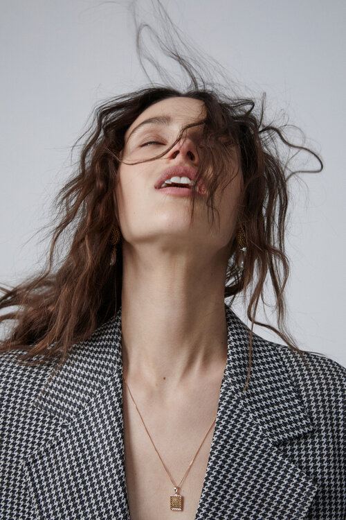 Portrait of a young brunette woman with wavy hair blowing, wearing a houndstooth blazer and a gold necklace against a light background.