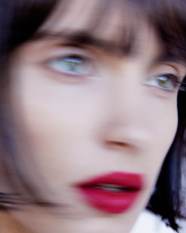 Close-up portrait of a young adult woman with short dark hair, red lipstick, and fair skin, looking off-camera with a blurred effect.