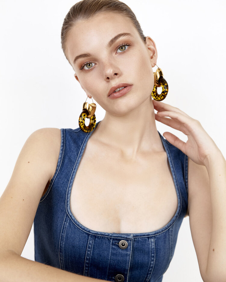 Close-up of a fair-skinned young woman in a denim top, posing in front of a white backdrop, showcasing gold and tortoiseshell earrings.