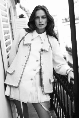 Model leaning on a balcony railing wearing a textured tweed jacket and white pleated skirt in a monochrome photograph.