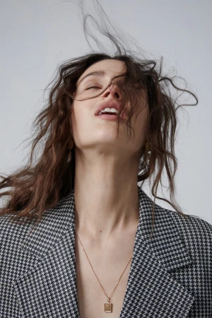 Close-up portrait of a woman with tousled hair wearing a houndstooth blazer and delicate gold pendant necklace.