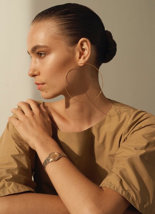 Elegant woman in a tan dress and gold hoop earrings poses in a minimalist indoor setting with soft, diffused natural lighting.