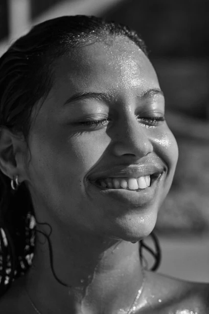 Black and white portrait of a young woman with wet skin and hair, smiling with closed eyes, wearing hoop earrings and a delicate necklace.