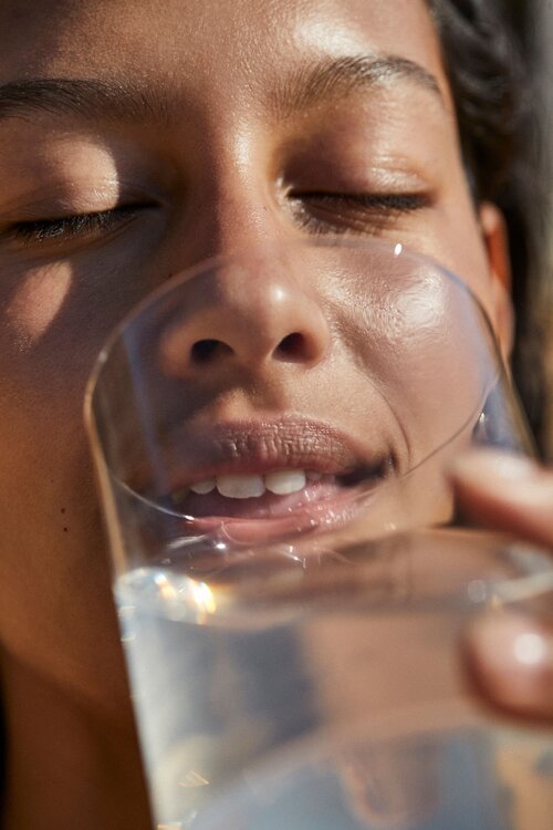 Close-up portrait of a young adult female holding a glass of water in front of her face, illuminated by natural light.