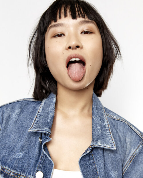 Close-up of a young Asian woman with short hair and a denim jacket, sticking out her tongue in a playful expression against a white background.