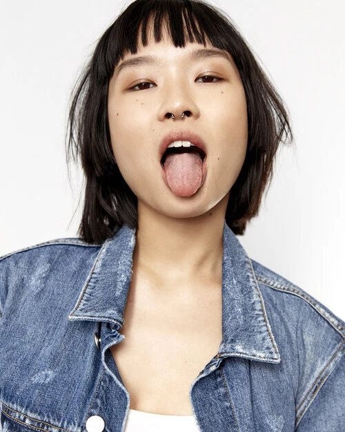 Close-up of a young Asian woman with short hair and a denim jacket, sticking out her tongue in a playful expression against a white background.