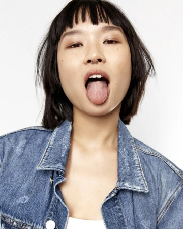 Close-up of a young Asian woman with short hair and a denim jacket, sticking out her tongue in a playful expression against a white background.