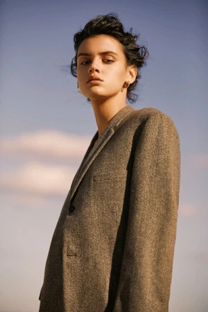 Portrait of a model wearing a textured brown blazer against a soft twilight sky, featuring makeup by Jasmin Lo.
