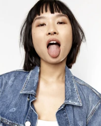 Close-up portrait of a woman with dark bob hair and septum piercing wearing a denim jacket sticking out her tongue