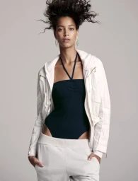 Model with voluminous dark hair wearing a dark halter top, white jacket, and light trousers against a neutral grey background.