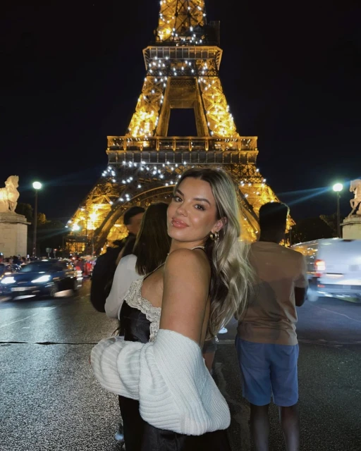 A woman poses in front of the Eiffel Tower at night, wearing a dress and sweater with city lights and people in the background.