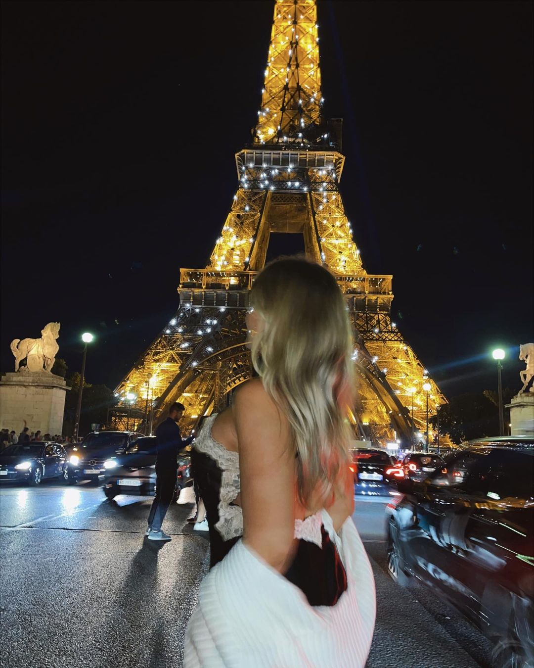 Woman gazes at the illuminated Eiffel Tower at night in Paris, with blurred cars and city lights adding to the vibrant urban scene.