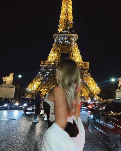 Woman gazes at the illuminated Eiffel Tower at night in Paris, with blurred cars and city lights adding to the vibrant urban scene.