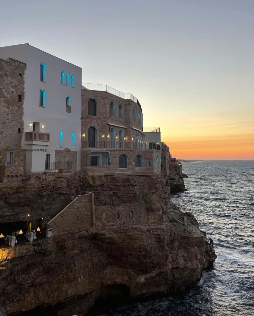 Coastal buildings sit on a rugged cliffside overlooking the sea at sunset, featuring stone architecture and illuminated windows.