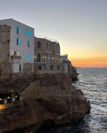 Coastal buildings sit on a rugged cliffside overlooking the sea at sunset, featuring stone architecture and illuminated windows.