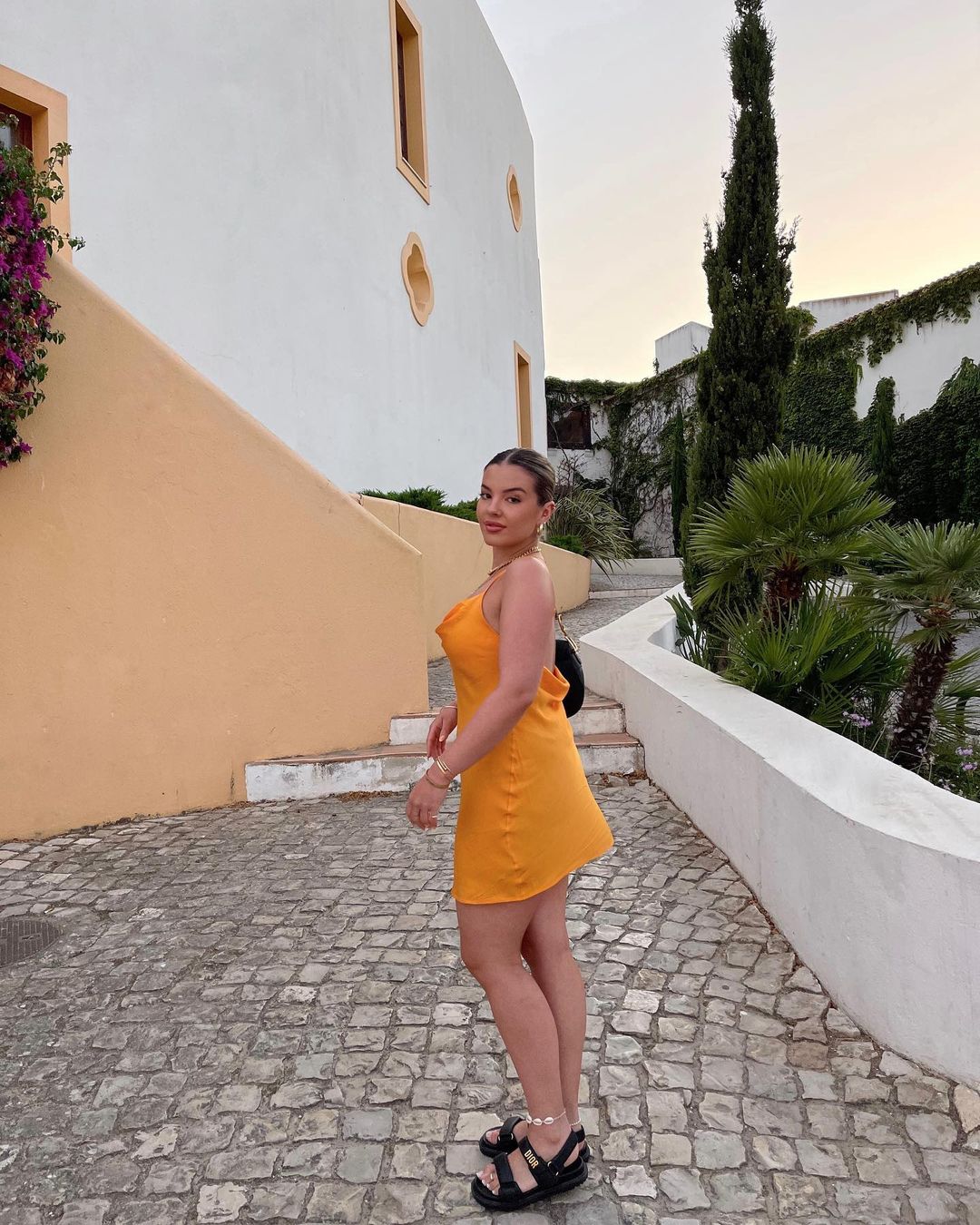 Woman in orange dress posing outdoors on cobblestone street with buildings and lush greenery in the background during daytime.