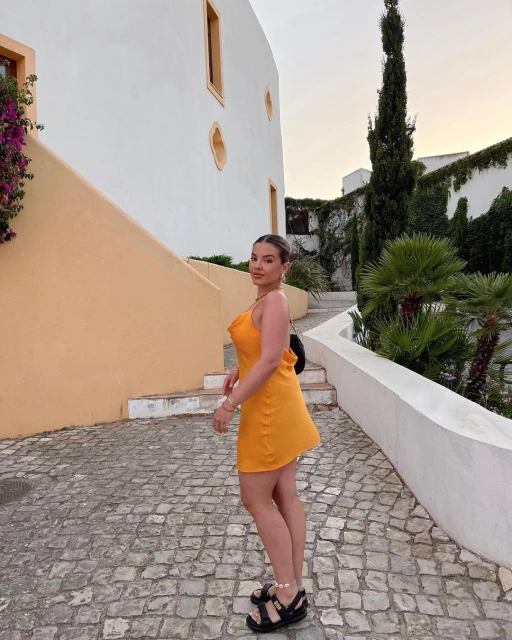 Woman in orange dress posing outdoors on cobblestone street with buildings and lush greenery in the background during daytime.