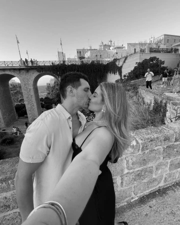 Monochrome selfie of a couple kissing on a stone bridge with historic buildings and archways in the background.
