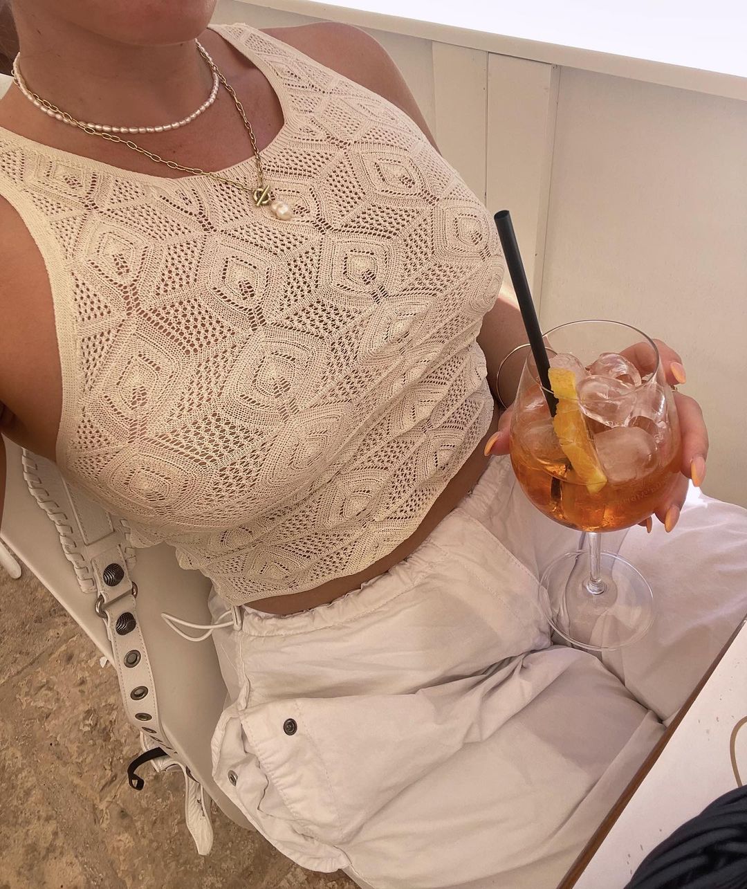 A person sitting at a table wearing a cream-colored knit tank top, with a drink in a wine glass, outdoors with natural light.