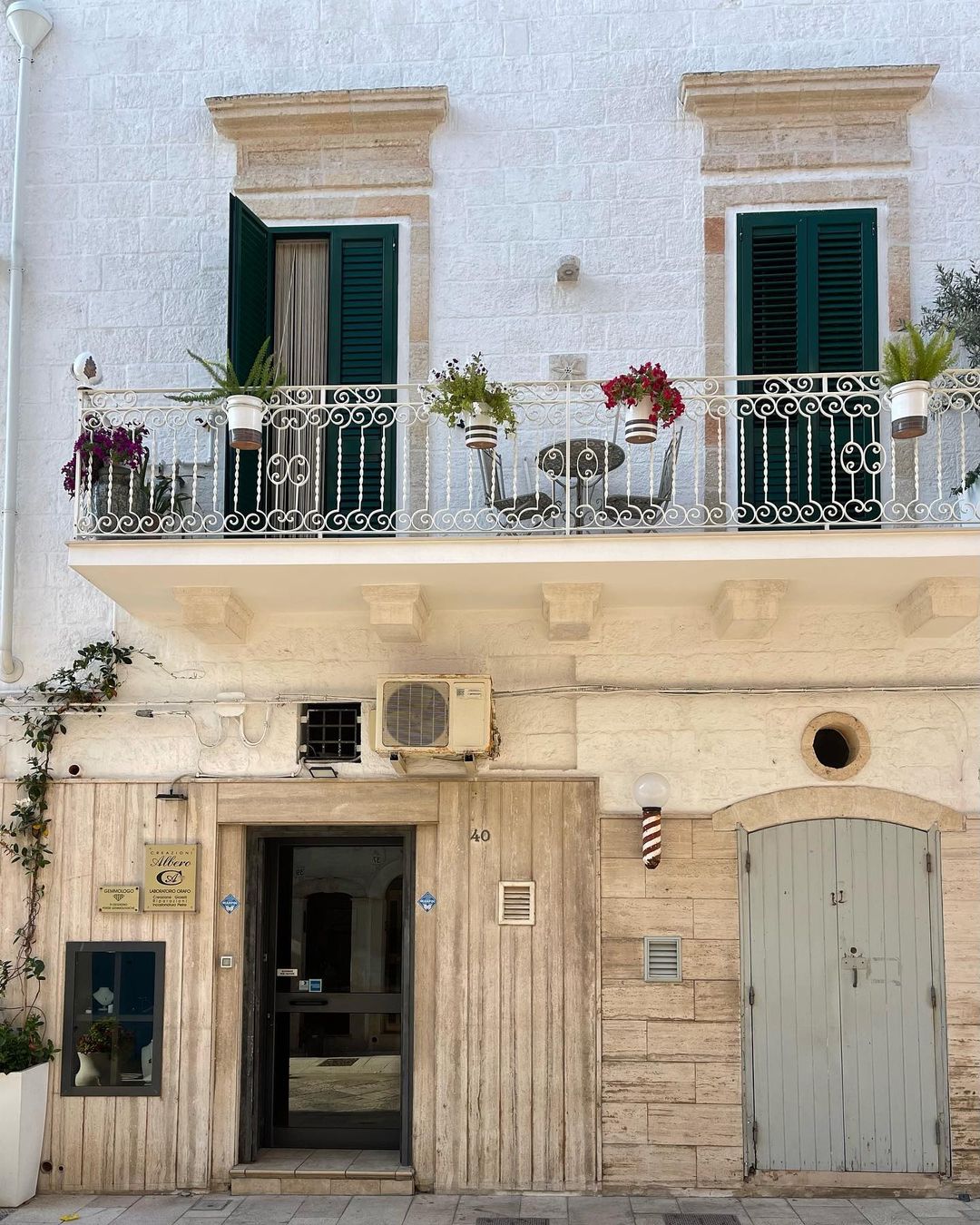 Exterior of a two-story building in Italy with a balcony, potted plants, white walls, dark green shutters, and a street-level entrance.