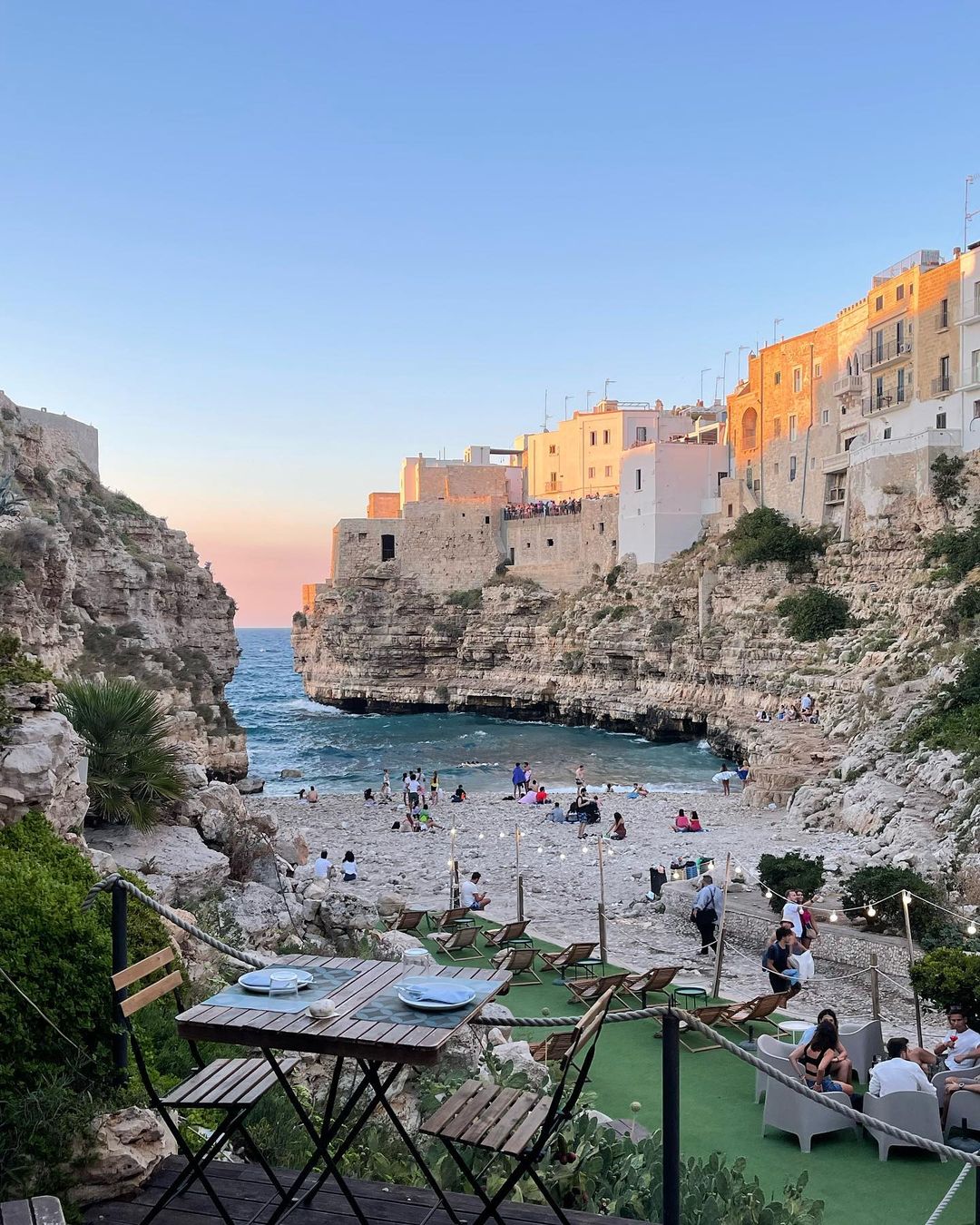 Cliffside beach scene in Polignano a Mare, Italy features buildings on cliffs, people on a beach, and a restaurant overlooking the water.