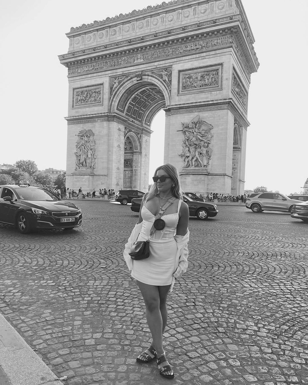 A woman poses in front of the Arc de Triomphe in Paris, wearing a white dress, sunglasses, and sandals on a cobblestone street.
