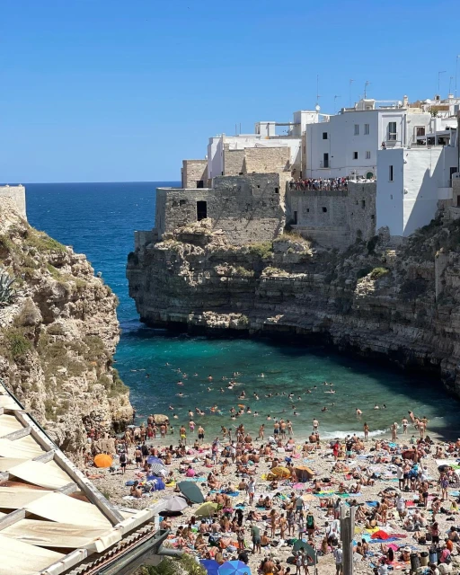 Crowded beach scene in Polignano a Mare, Italy, featuring numerous people enjoying the clear turquoise water and sandy shore.