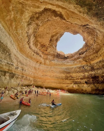 Dramatic view of Benagil Sea Cave with sandy beach, blue-green water, people kayaking, and natural sunlight creating a scenic seascape.