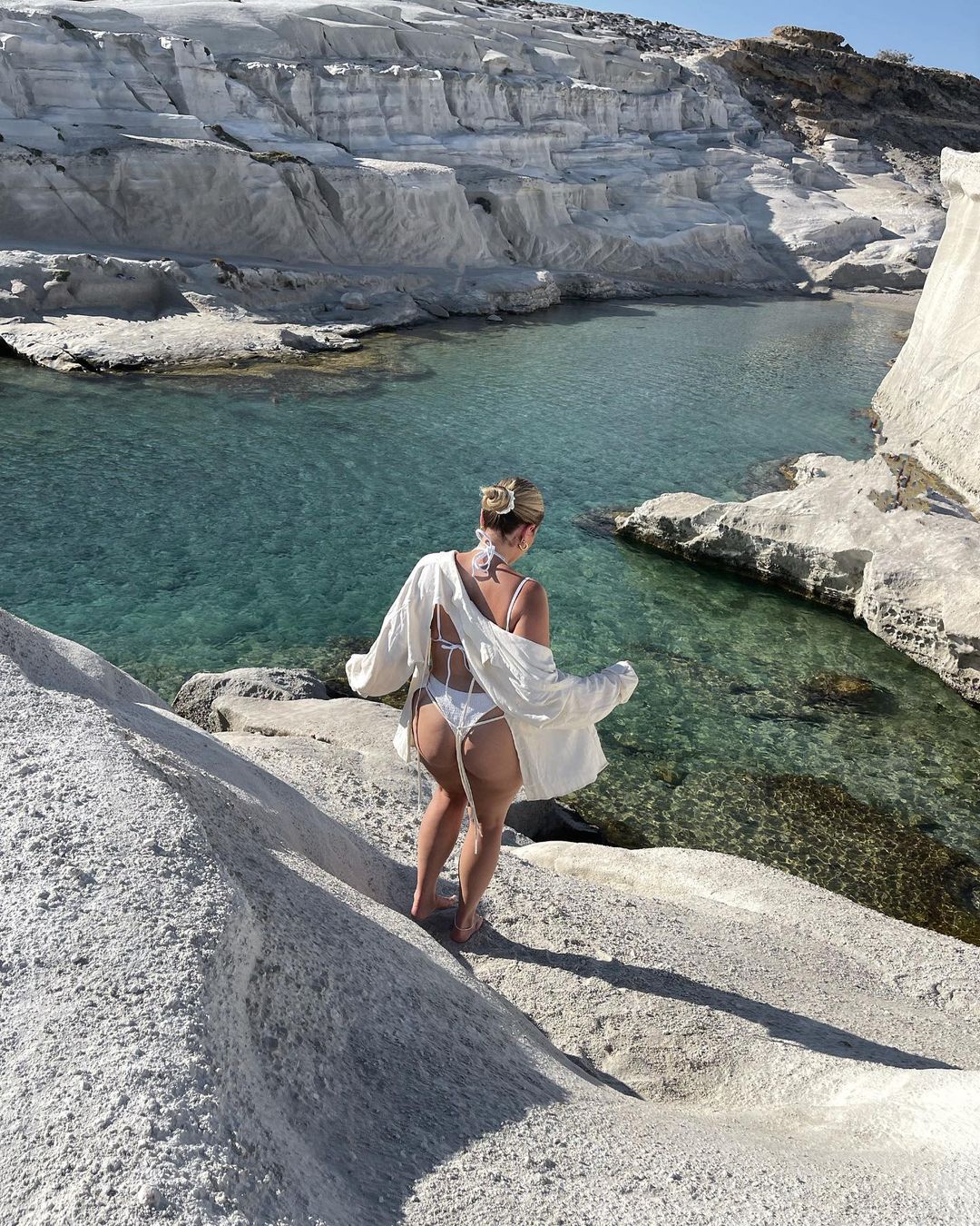 Woman in white swimwear and cover-up poses on a white rock formation overlooking clear water on a sunny day near a rock cliff.