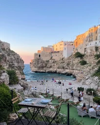 Coastal beach scene viewed from a cliffside terrace with dining tables, overlooking rocky cliffs and white buildings at sunset.