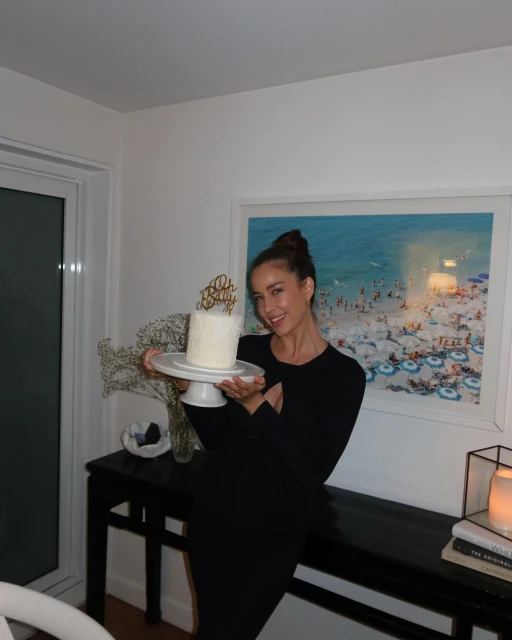 Adult woman in black dress smiles while holding up a cake on a stand in an indoor setting with a seascape painting visible.