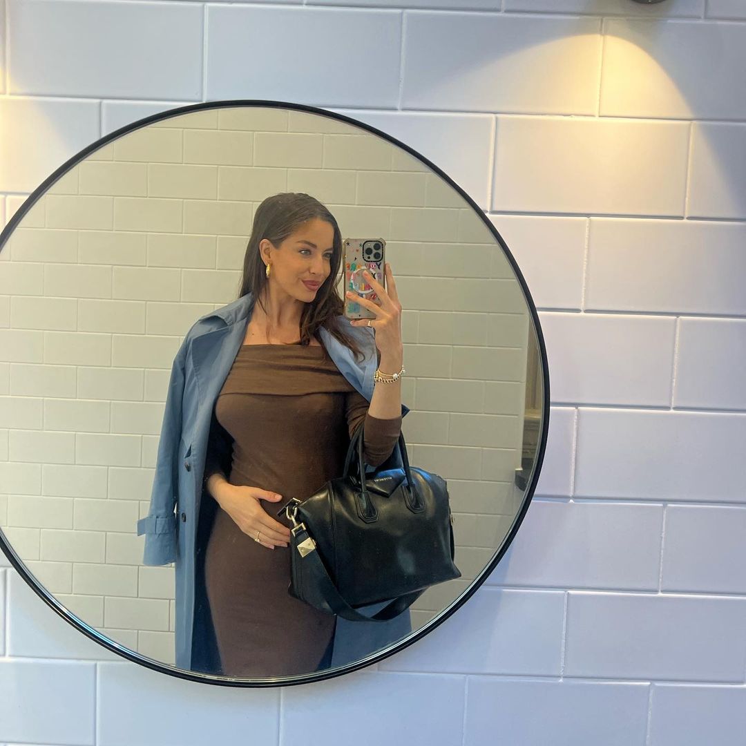 A woman in a brown dress takes a mirror selfie in a tiled bathroom, wearing a blue coat and holding a black leather handbag.