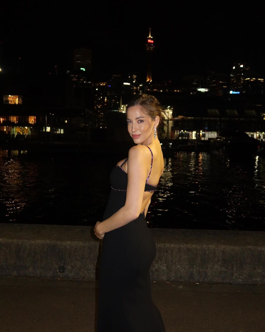 A woman in a black dress poses at night with a cityscape and water in the background, creating a captivating evening portrait.