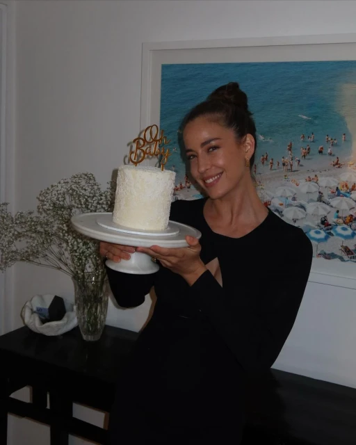 Woman holds birthday cake in indoor setting. Blonde female smiles near cake decorated with a golden topper in domestic environment.