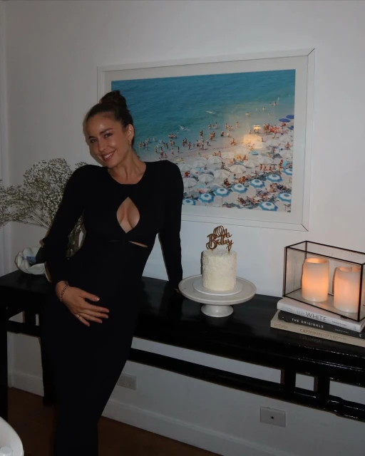 Adult woman in a black dress poses with a cake near a black table with decorative objects in an indoor environment with beach art.