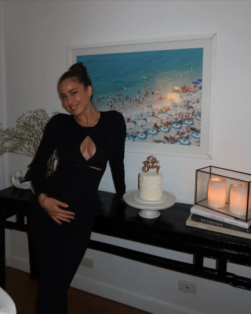 Adult woman in a black dress poses with a cake near a black table with decorative objects in an indoor environment with beach art.