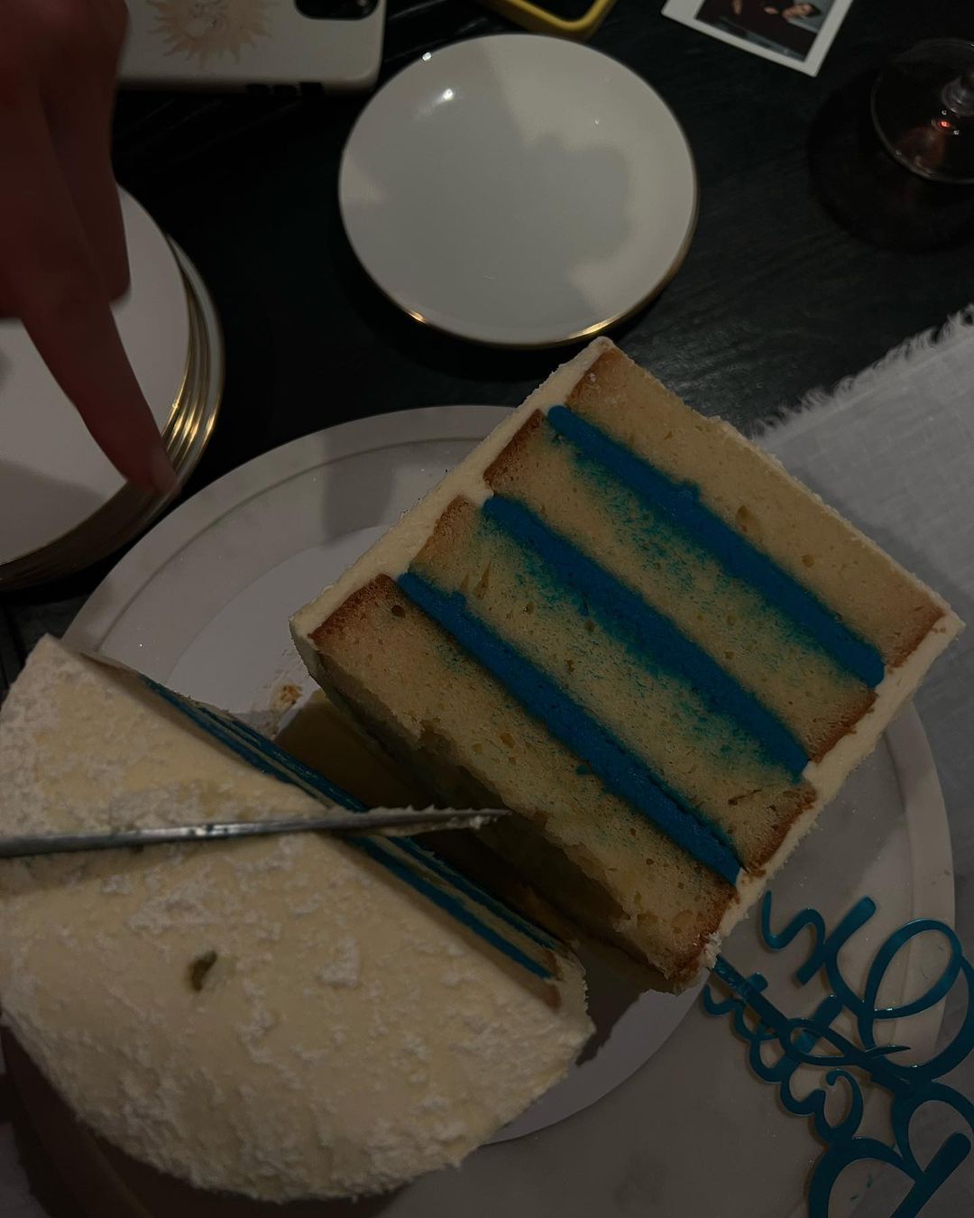 A slice being cut from a multi-layered cake with white frosting and blue filling on a white plate at an indoor setting.
