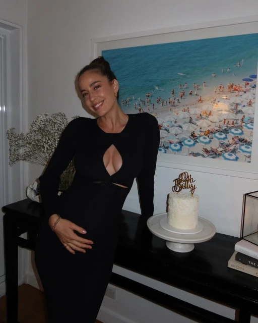 Pregnant woman in black dress poses indoors with cake in front of a beach scene photograph, celebrating baby to be.