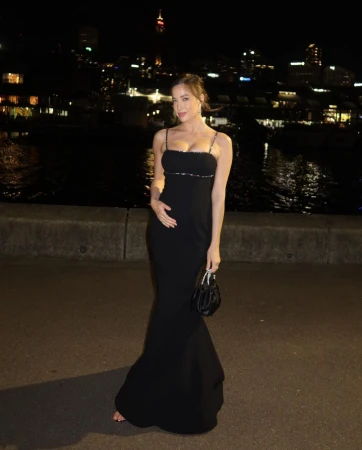 Woman in a long black dress holding a small black handbag against a night waterfront backdrop.