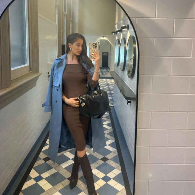 Adult female in brown dress and boots takes mirror selfie indoors, wearing a blue trench coat, and carrying a large black handbag.