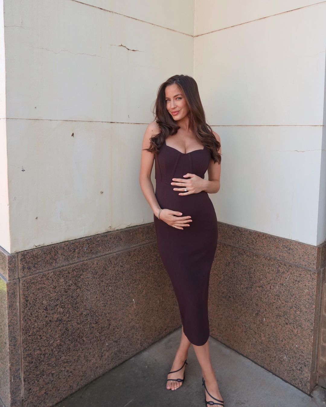 A pregnant woman with long dark hair poses in a brown dress and strappy sandals, holding her stomach in front of a neutral background.