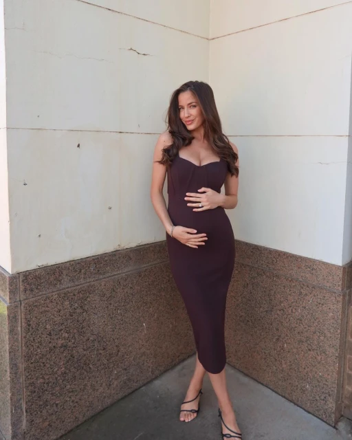A pregnant woman with long dark hair poses in a brown dress and strappy sandals, holding her stomach in front of a neutral background.